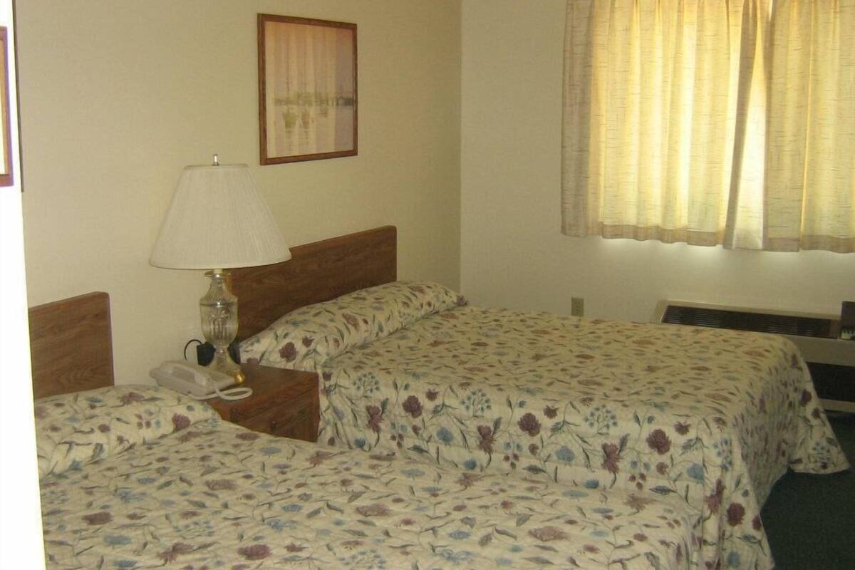 A hotel room with two beds, a bedside table with a lamp, a framed picture on the wall, and a window with curtains.