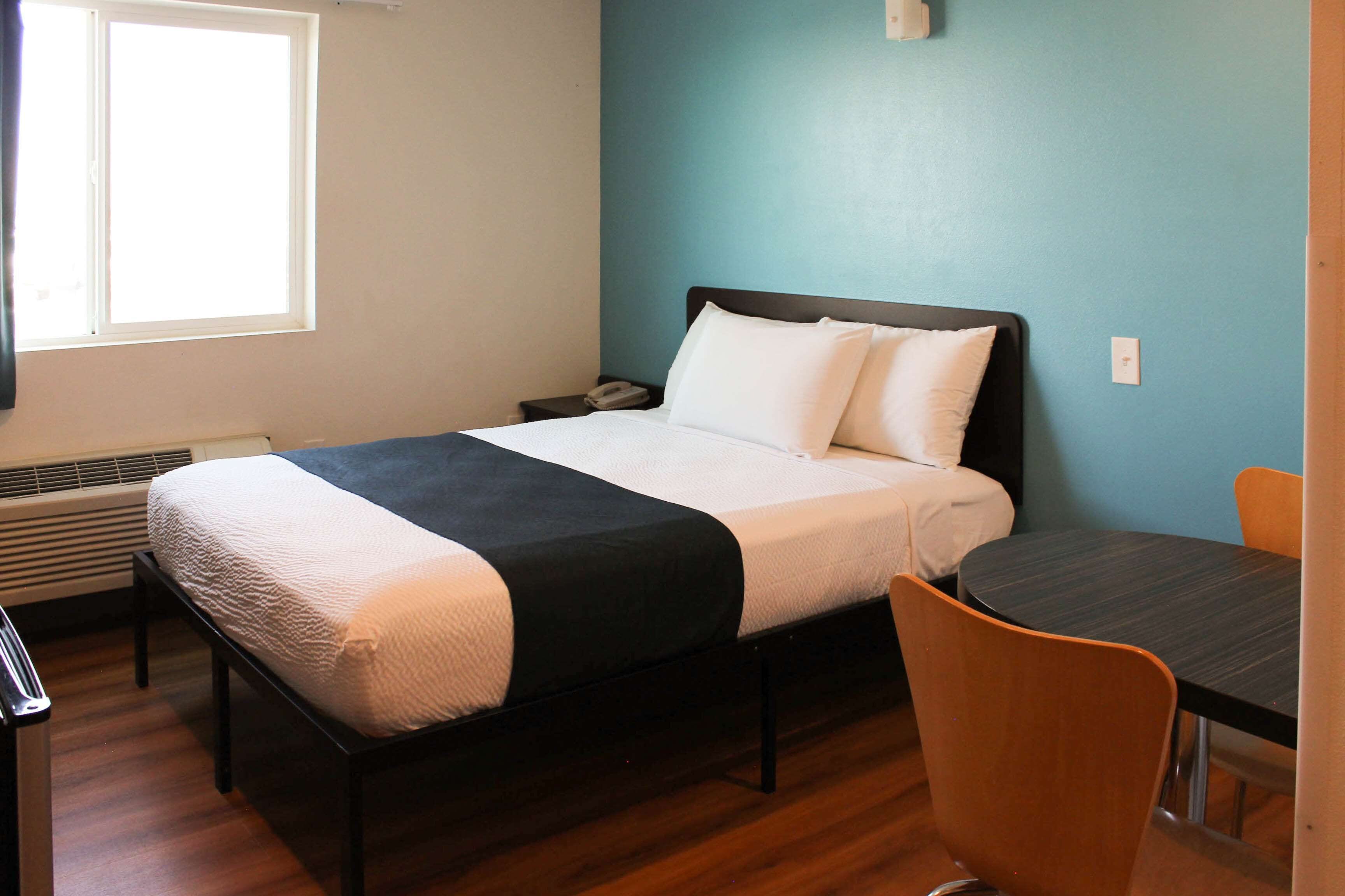 Una cama individual con estructura negra, ropa de cama blanca y una manta negra. Hay una mesita de noche pequeña con un teléfono y una lámpara fijada a la pared. Una silla está colocada junto a una pequeña mesa cerca de la cama.