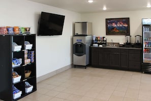 A hotel lobby with a flat-screen TV, a water dispenser, a coffee machine, a refrigerator stocked with beverages, and a snack bar with various packaged goods.