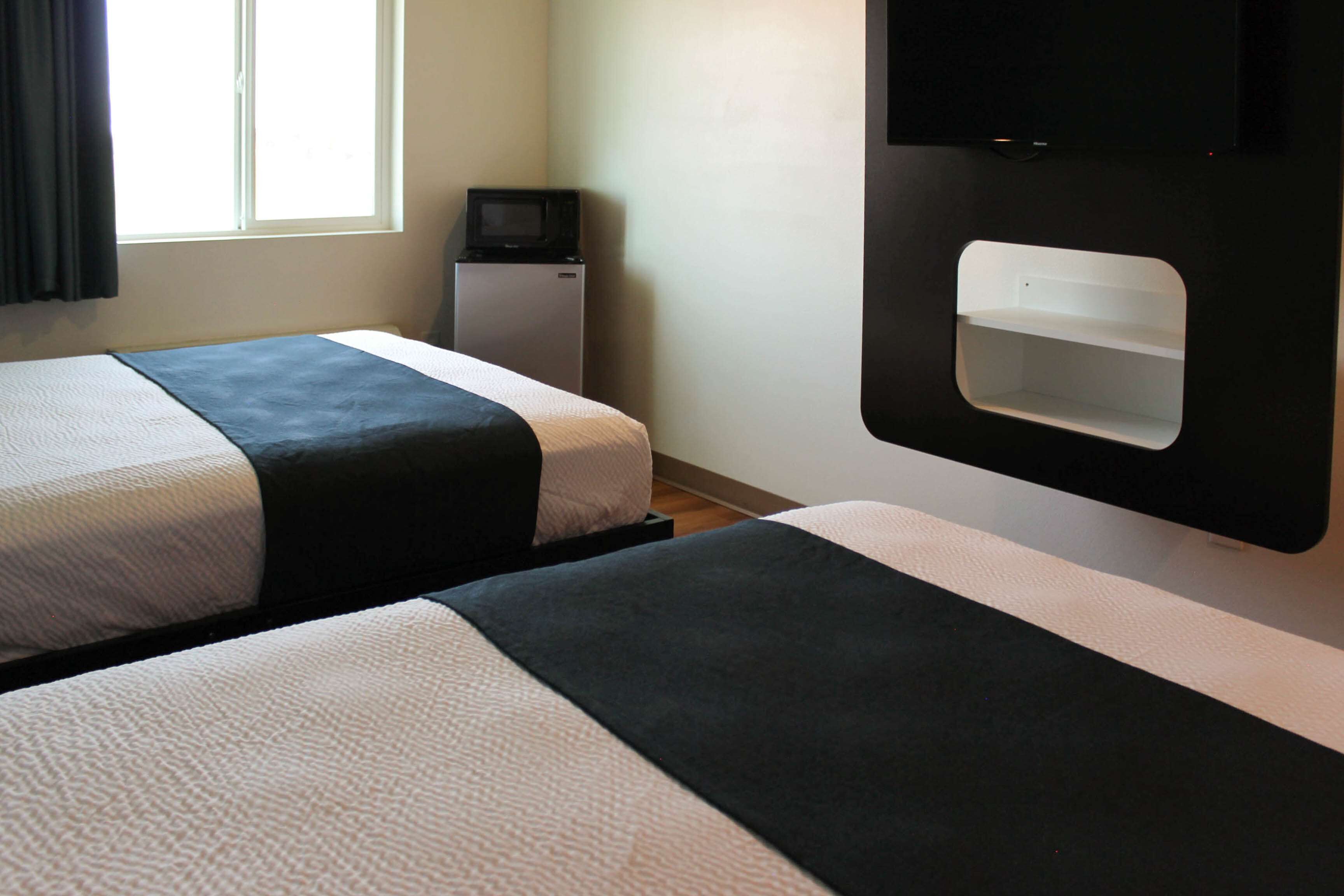 A hotel room with two beds, a small refrigerator, a TV mounted on the wall, and a window with curtains.