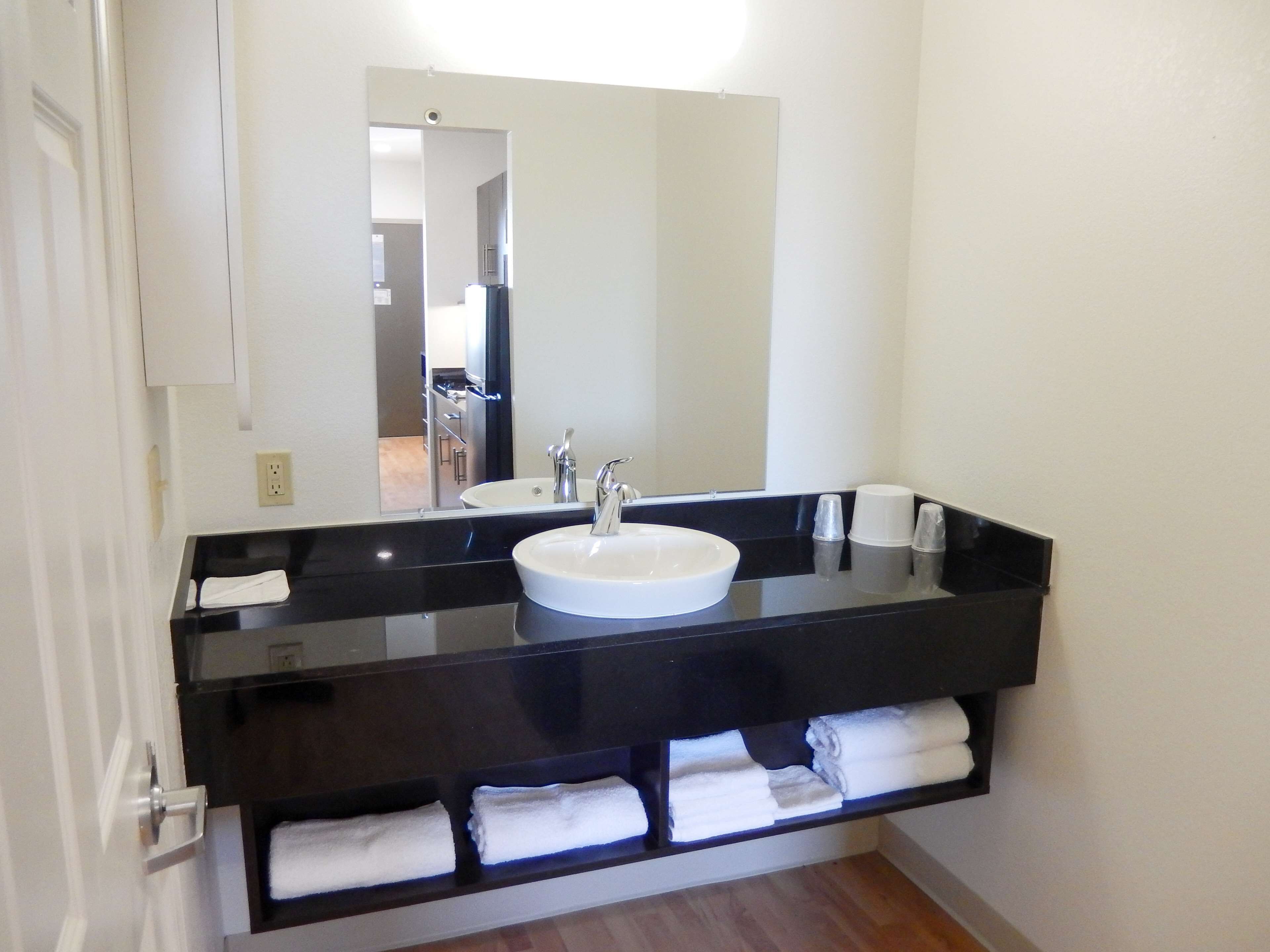 Un baño con encimera negra, lavabo blanco y un espejo. Hay toallas en un estante debajo del lavabo.