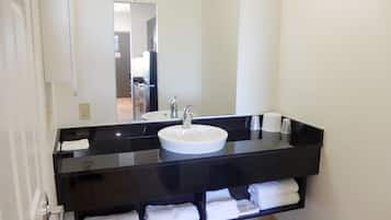 Un baño con encimera negra, lavabo blanco y un espejo. Hay toallas en un estante debajo del lavabo.