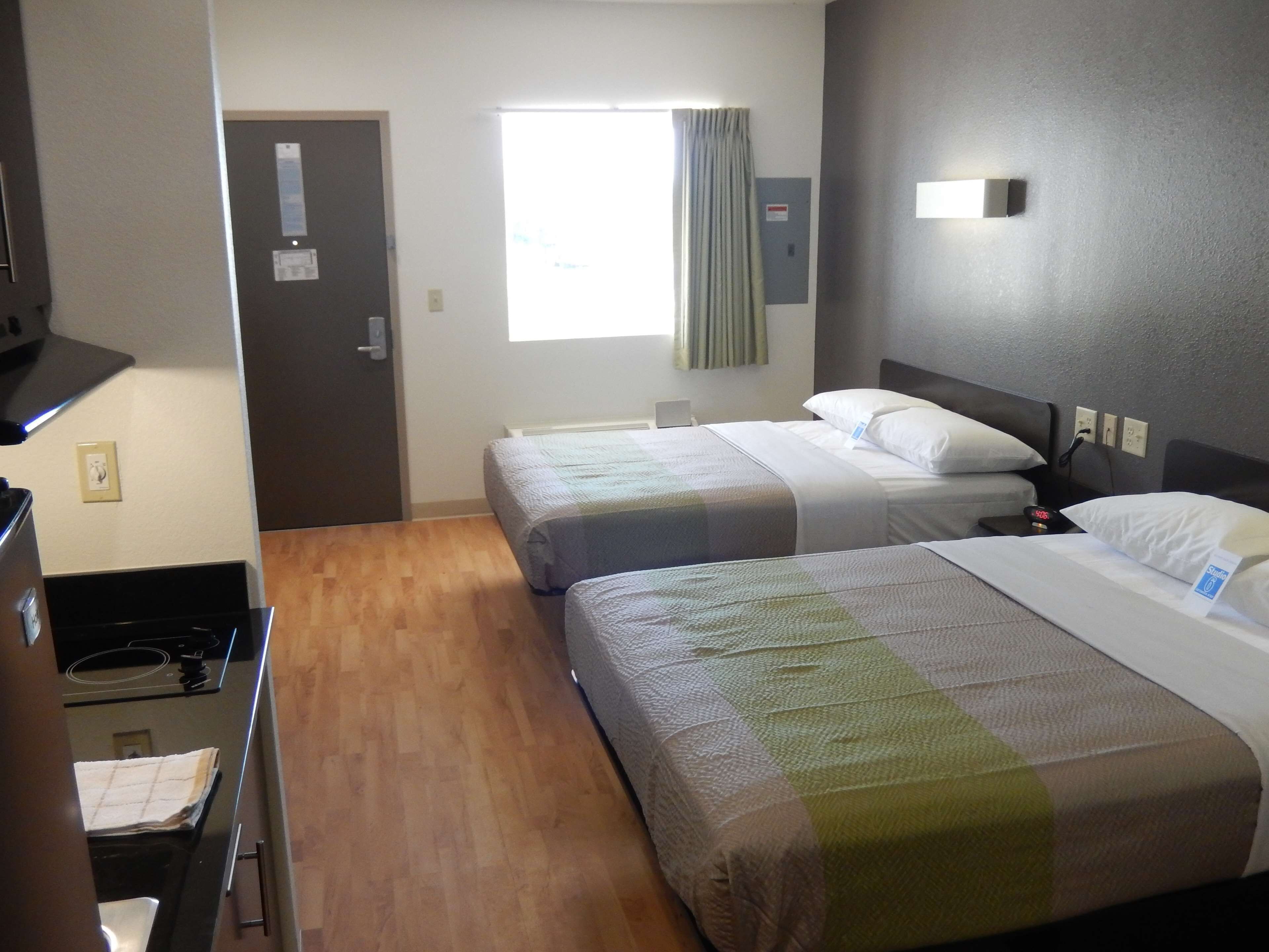 Ein Hotelzimmer mit zwei Betten, einer Küchenzeile, einem Fenster mit Vorhängen und einer Wandleuchte.