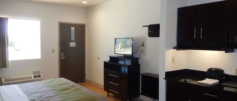 Habitación de hotel con cama, escritorio, televisión, kitchenette y ventana con cortinas.