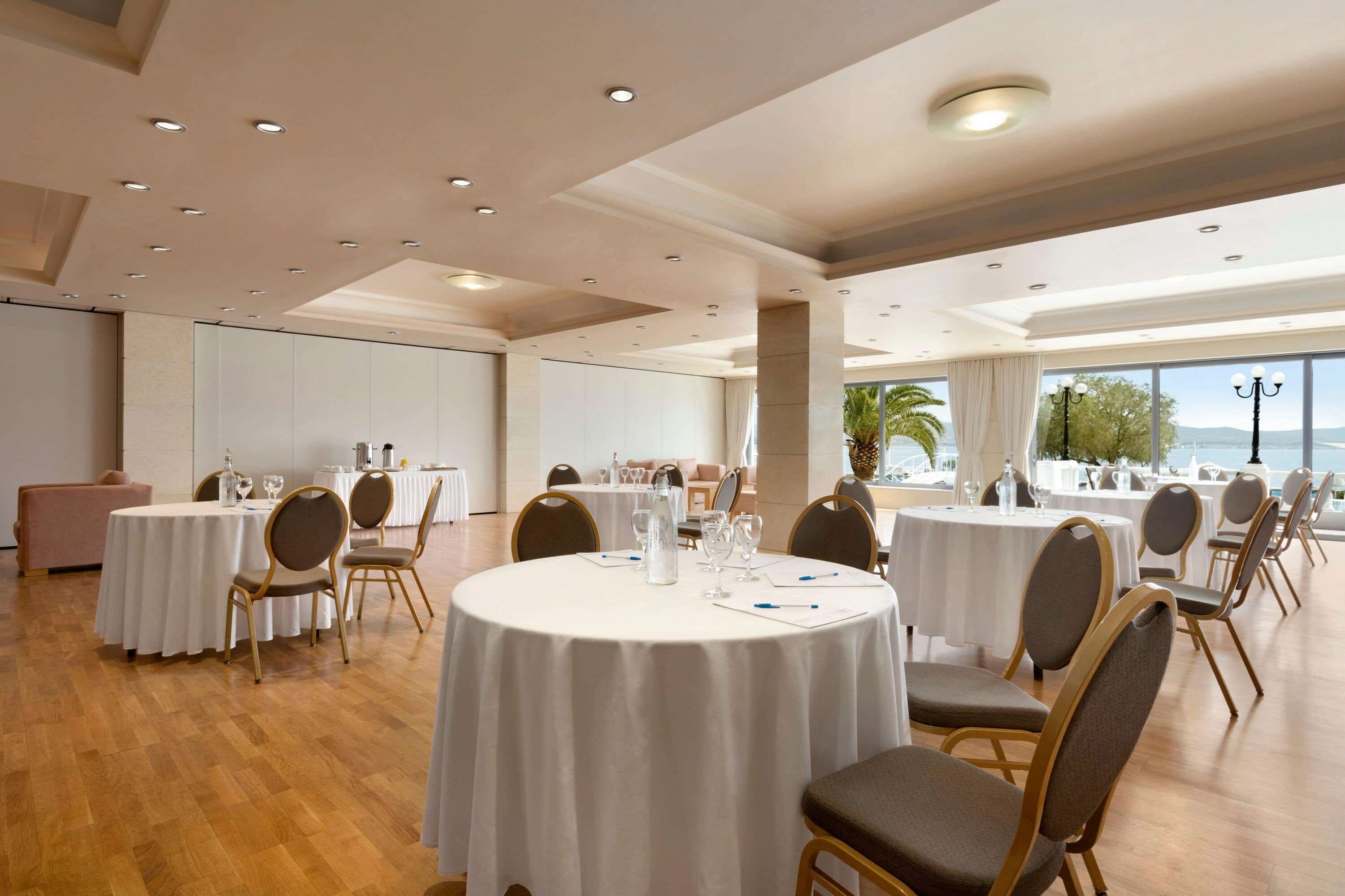 A spacious banquet hall with round tables and chairs, large windows offering a view of the sea, and a high ceiling with recessed lighting.