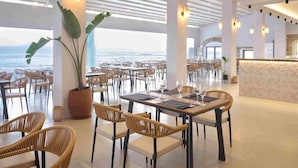 A spacious dining area with tables set for two, large potted plants, and a view of the ocean.