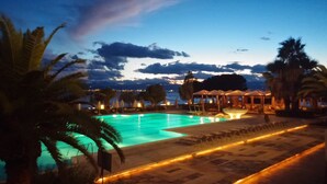 A swimming pool with lounge chairs and umbrellas, surrounded by palm trees and a view of the sea at dusk.