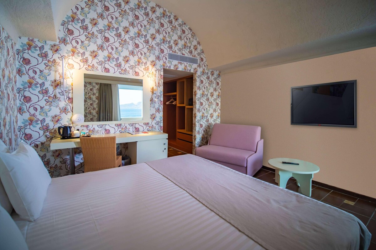 A hotel room with a bed, a desk with a chair, a pink chair, a small table, a wardrobe, and a TV on the wall.