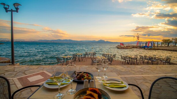 A table set with plates of food and wine glasses, overlooking a body of water with a diving boat and a pier in the background.