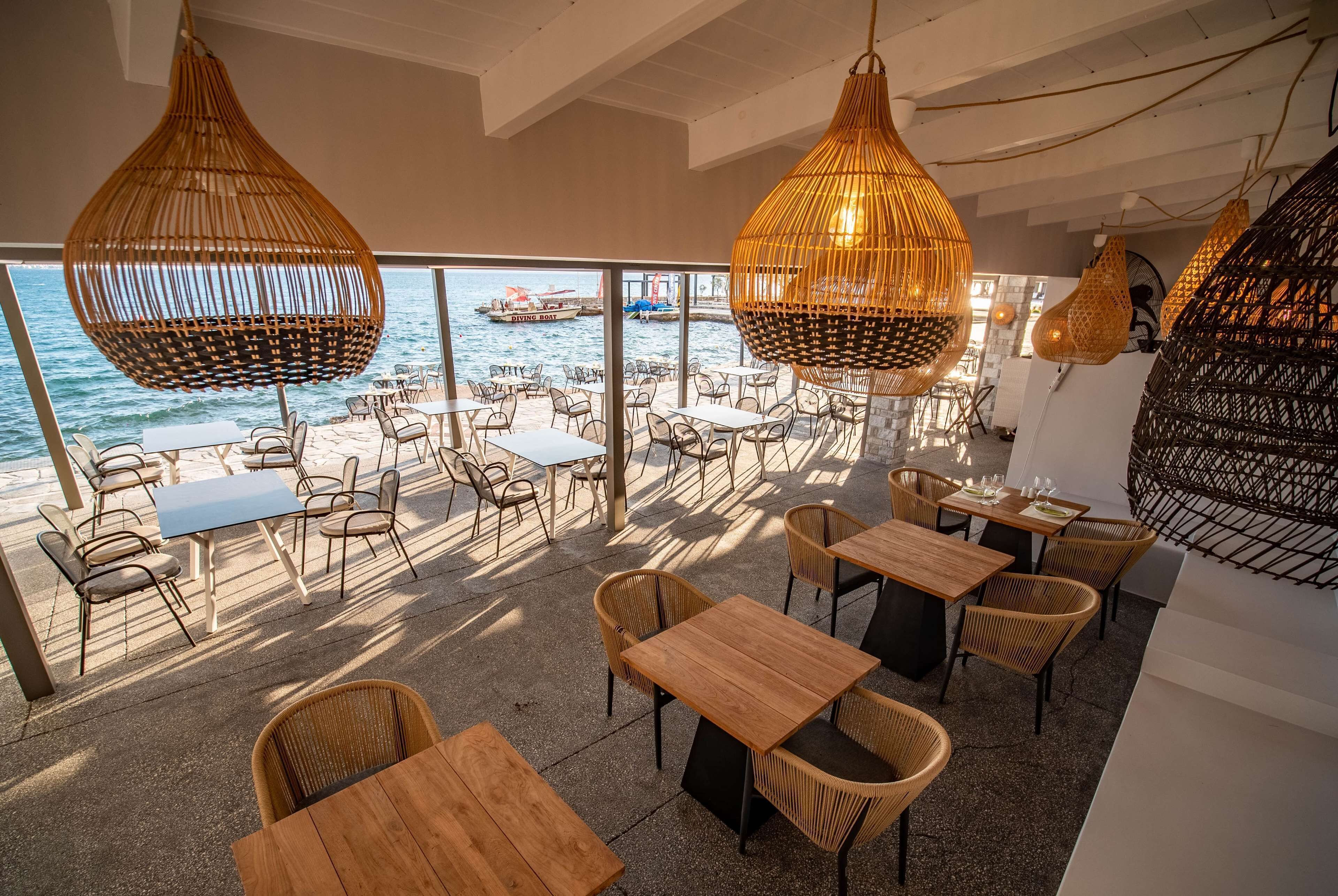 An outdoor dining area with wicker furniture and tables, overlooking the sea with boats visible in the background.