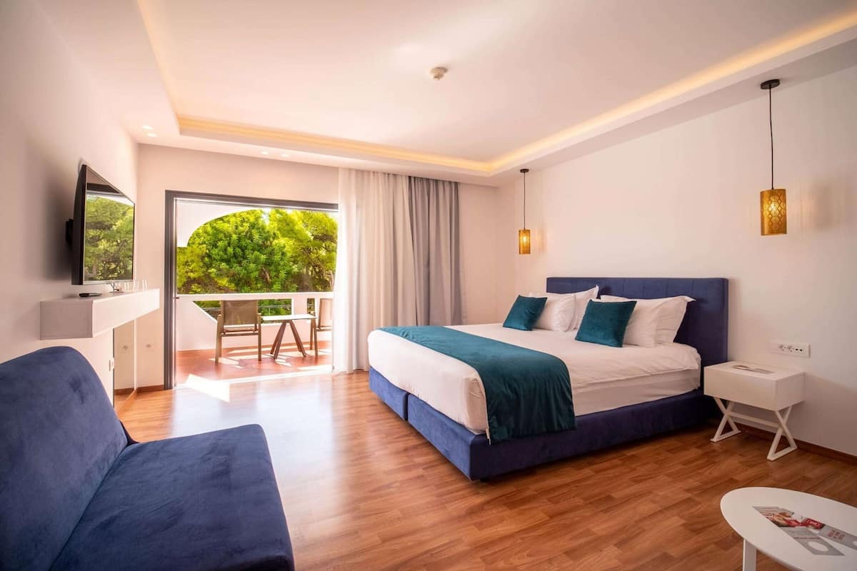 Ein modernes Hotelzimmer mit einem großen Bett, einer Sitzecke, einem kleinen Tisch und einem Balkon mit Blick ins Grüne.