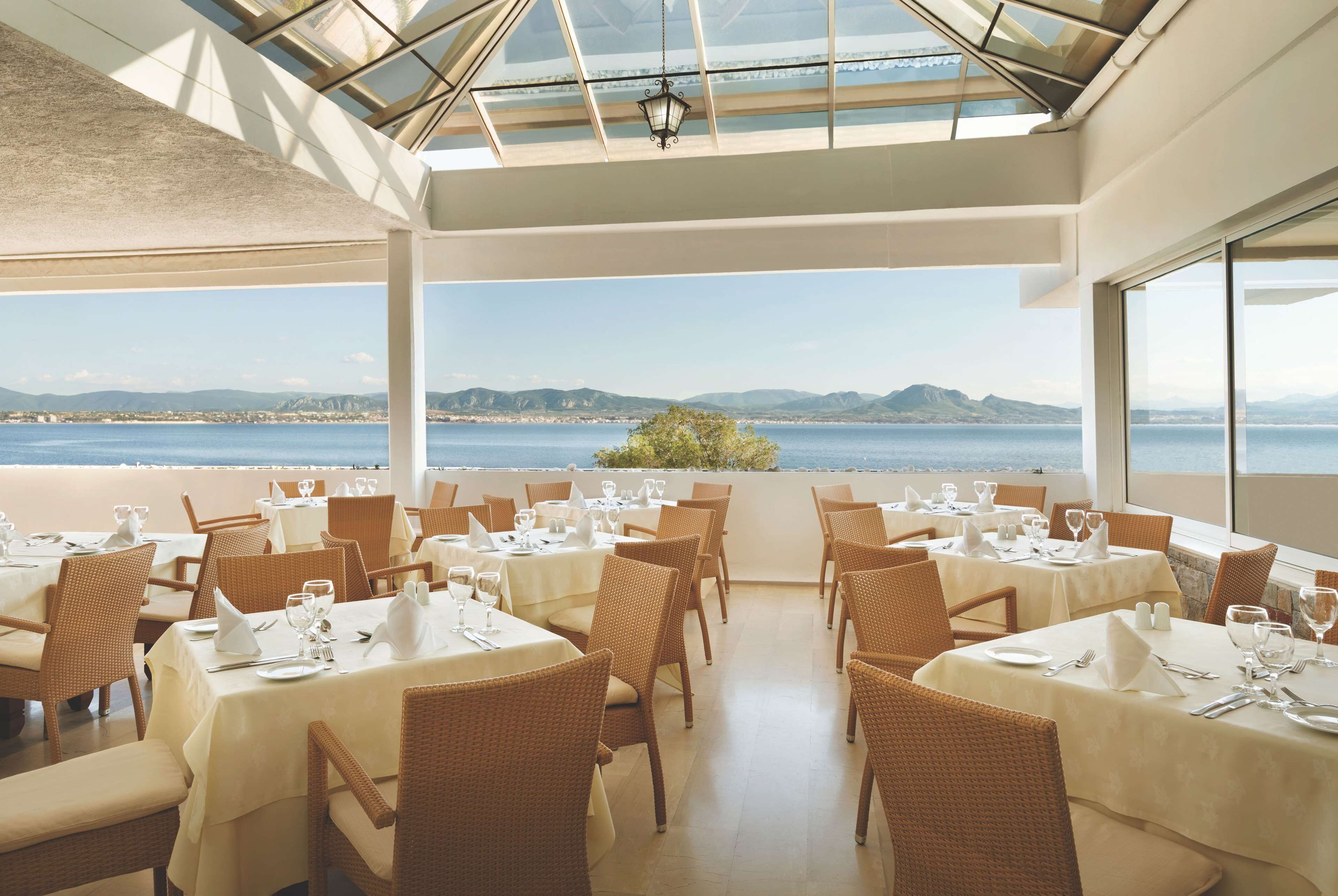 Un restaurant dont les tables sont dressées pour le service, offrant une vue panoramique sur la mer et les collines.