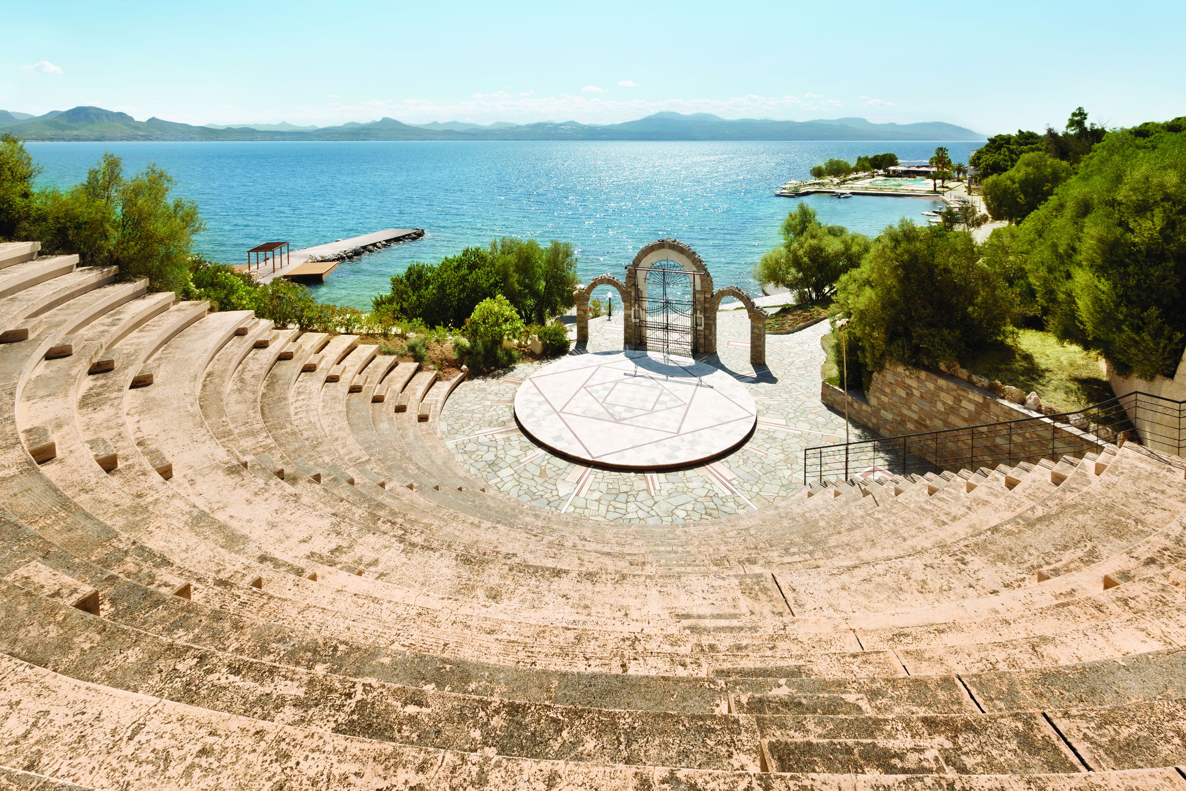 Um anfiteatro ao ar livre com palco central, degraus de pedra e vista para o mar.