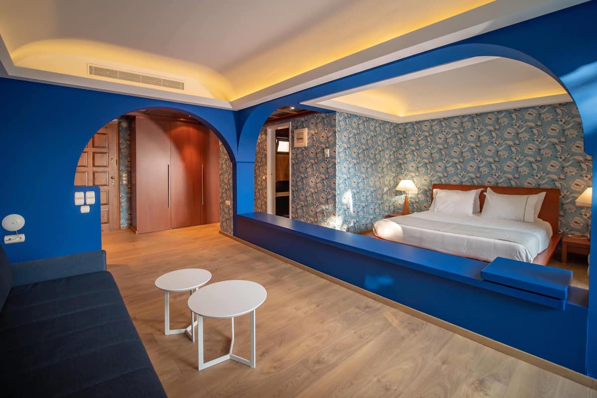 Ein modernes Hotelzimmer mit einem großen Bett, einem Schreibtisch und zwei Stühlen. Eine Holztür und eine blaue Wand mit floralem Muster sind ebenfalls zu sehen.