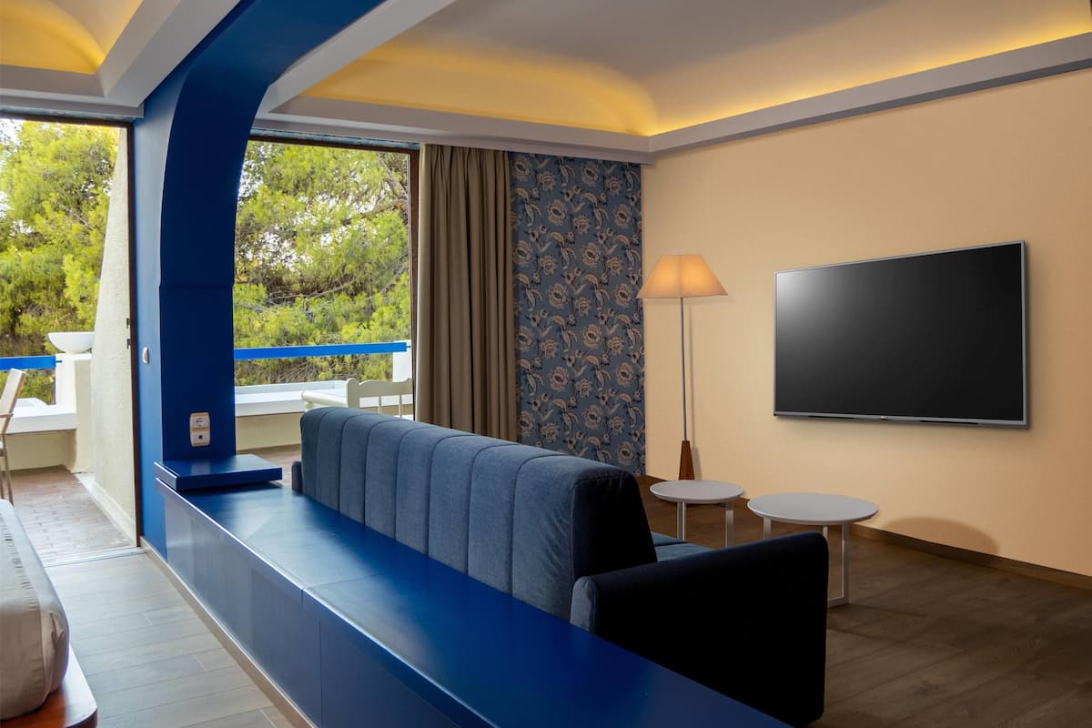 Ein modernes Hotelzimmer mit einem blauen Sofa, einem Flachbildfernseher und einem Balkon mit Blick auf Bäume.
