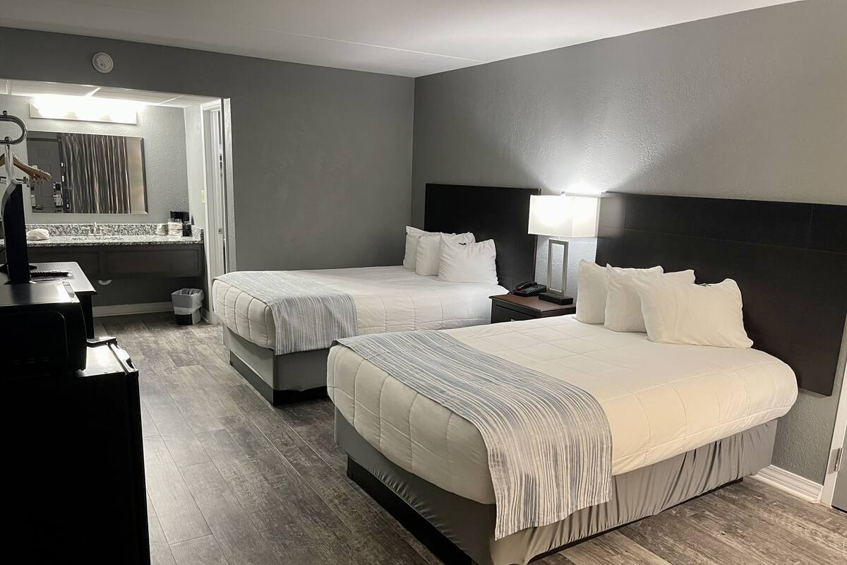 A hotel room with two beds, a desk, a mirror, and a bathroom visible through an open door.