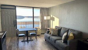 Una sala de estar con un sofá gris, una mesa de comedor con sillas, un ventilador de techo y vistas a un lago a través de un ventanal corredero.