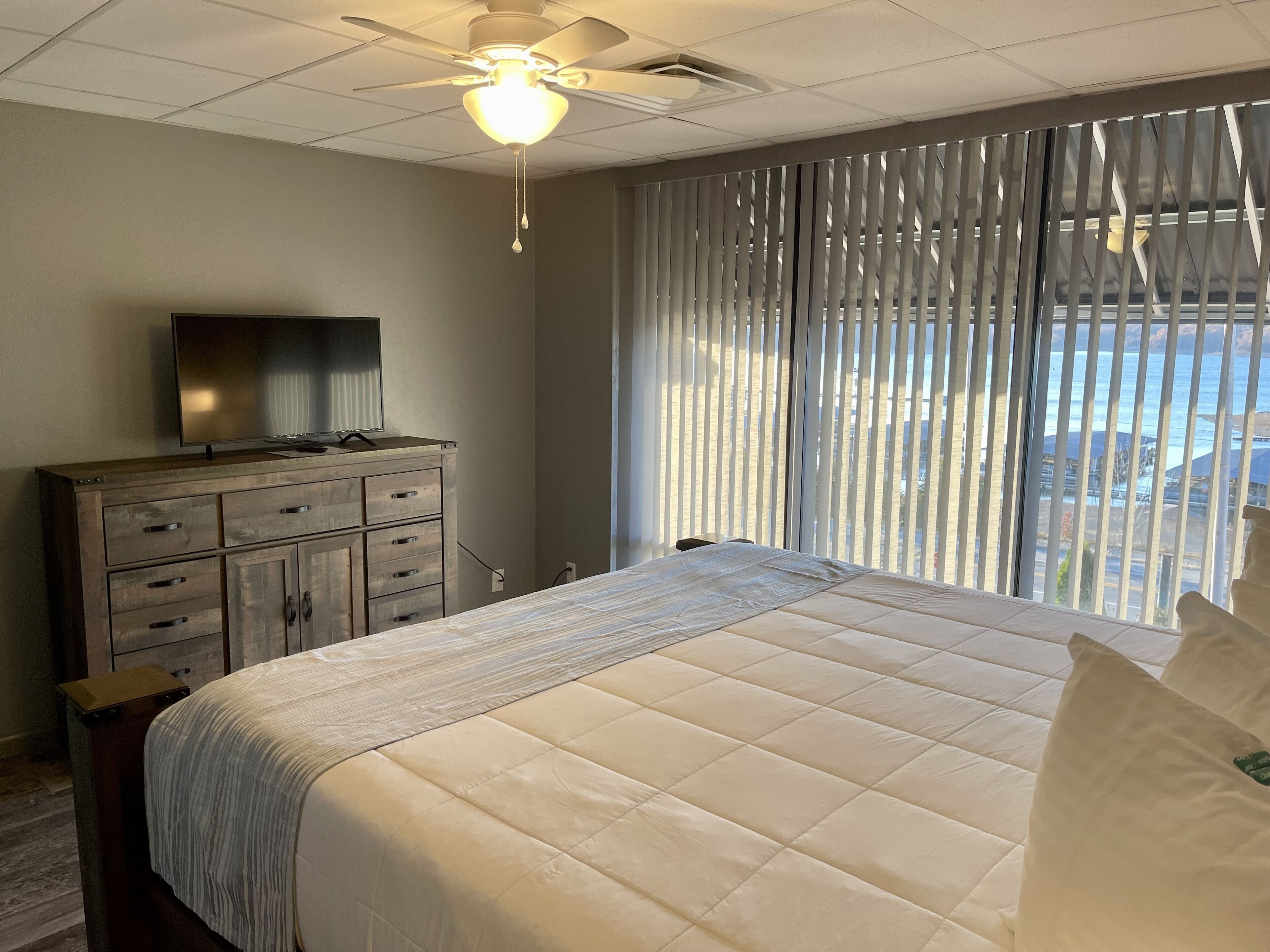 A bedroom with a bed, a TV on a dresser, a ceiling fan, and a view of the ocean through large windows.