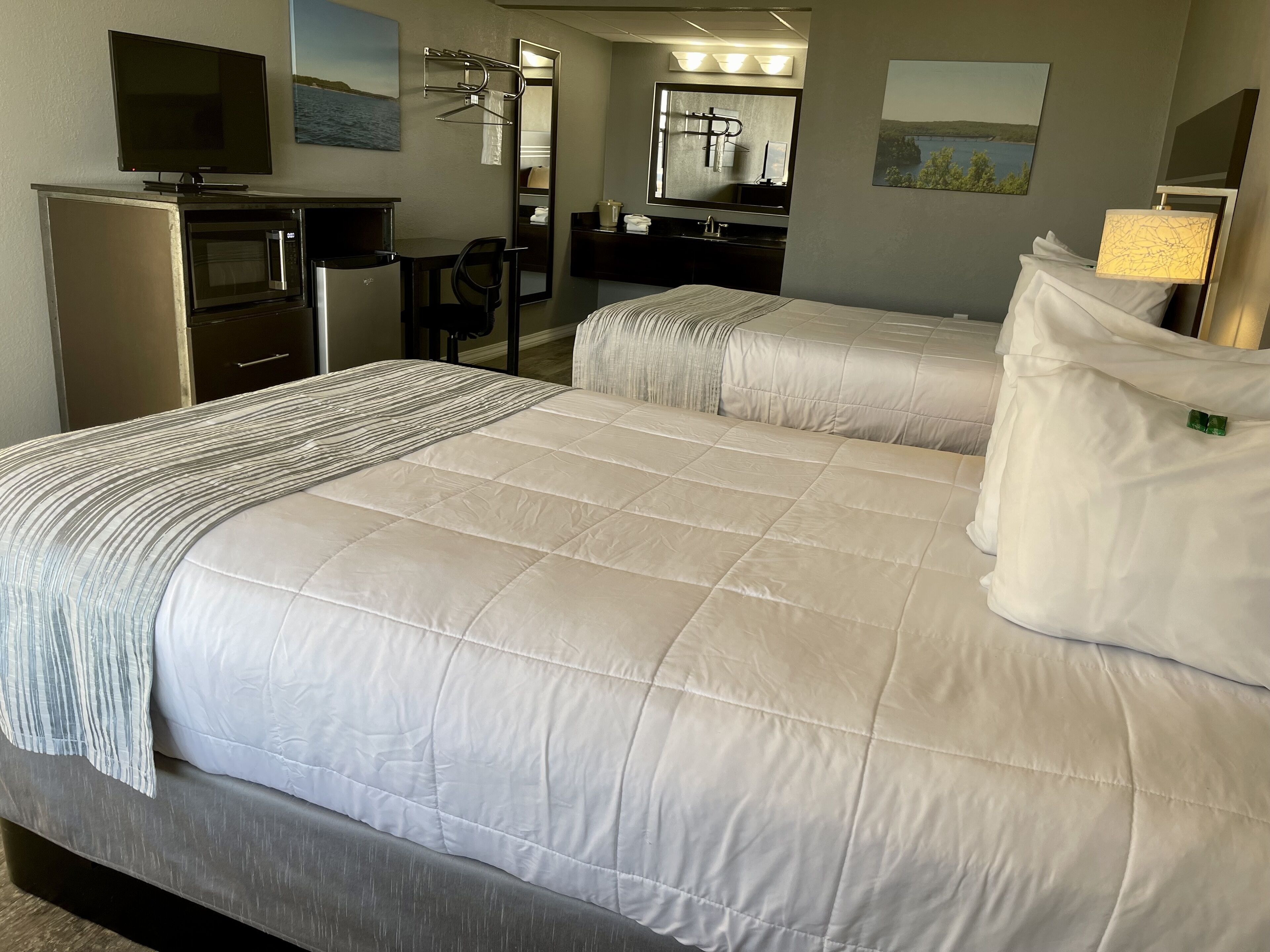 Una habitación de hotel con una cama grande, un escritorio con microondas, un televisor y un baño con espejo y perchero para toallas.