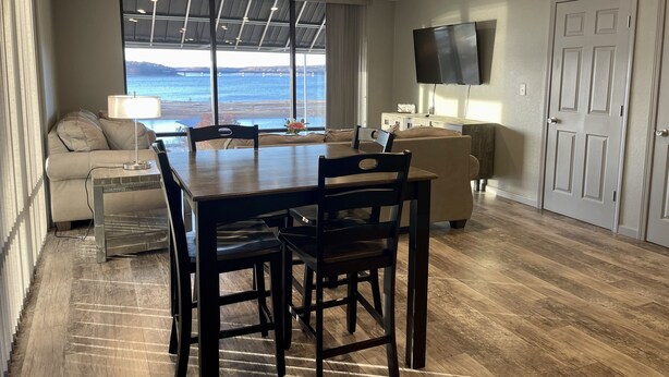 Un área de comedor con una mesa y sillas, un sofá, un televisor montado en la pared y vistas al agua a través de amplias ventanas.