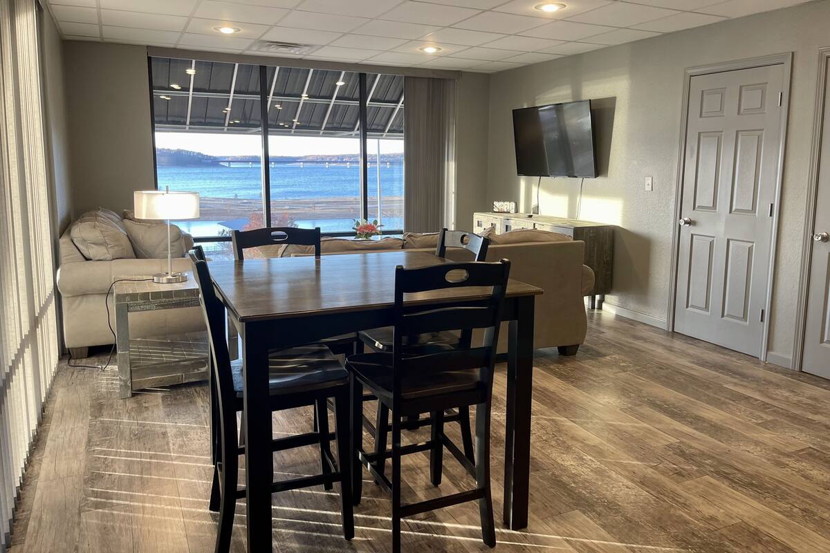 A dining area with a table and chairs, a sofa, a TV mounted on the wall, and a view of the water through large windows.