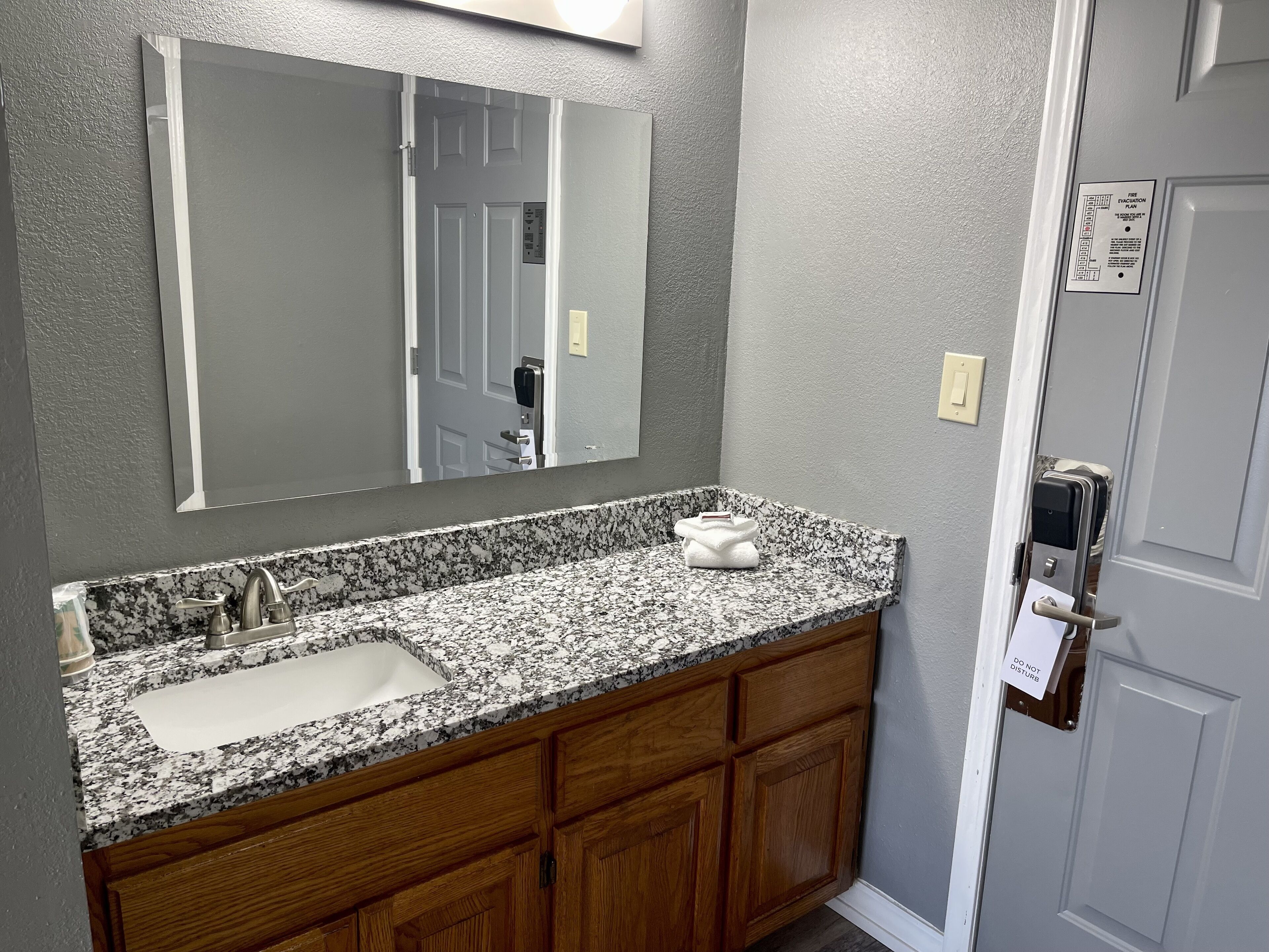 A bathroom with a granite countertop sink, wooden cabinets, a large mirror, and a door with a keypad lock.