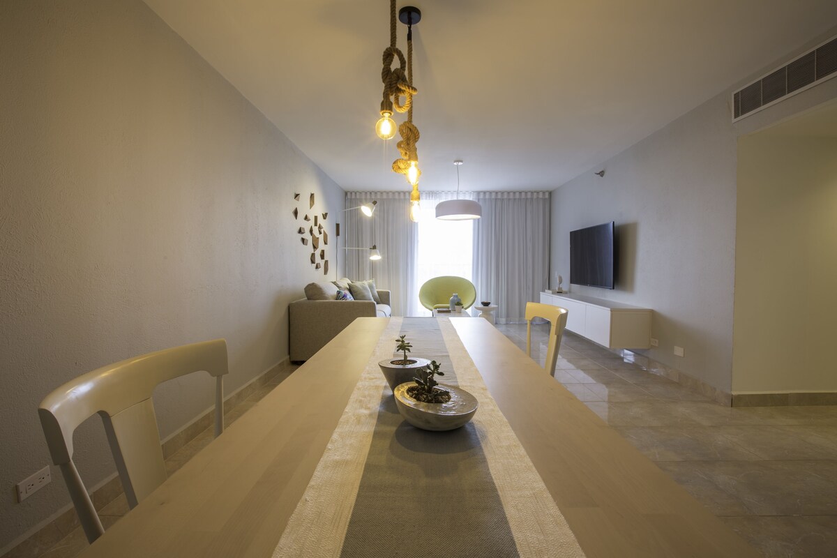 Un comedor moderno con una mesa larga, sillas, un adorno central e iluminación colgante.