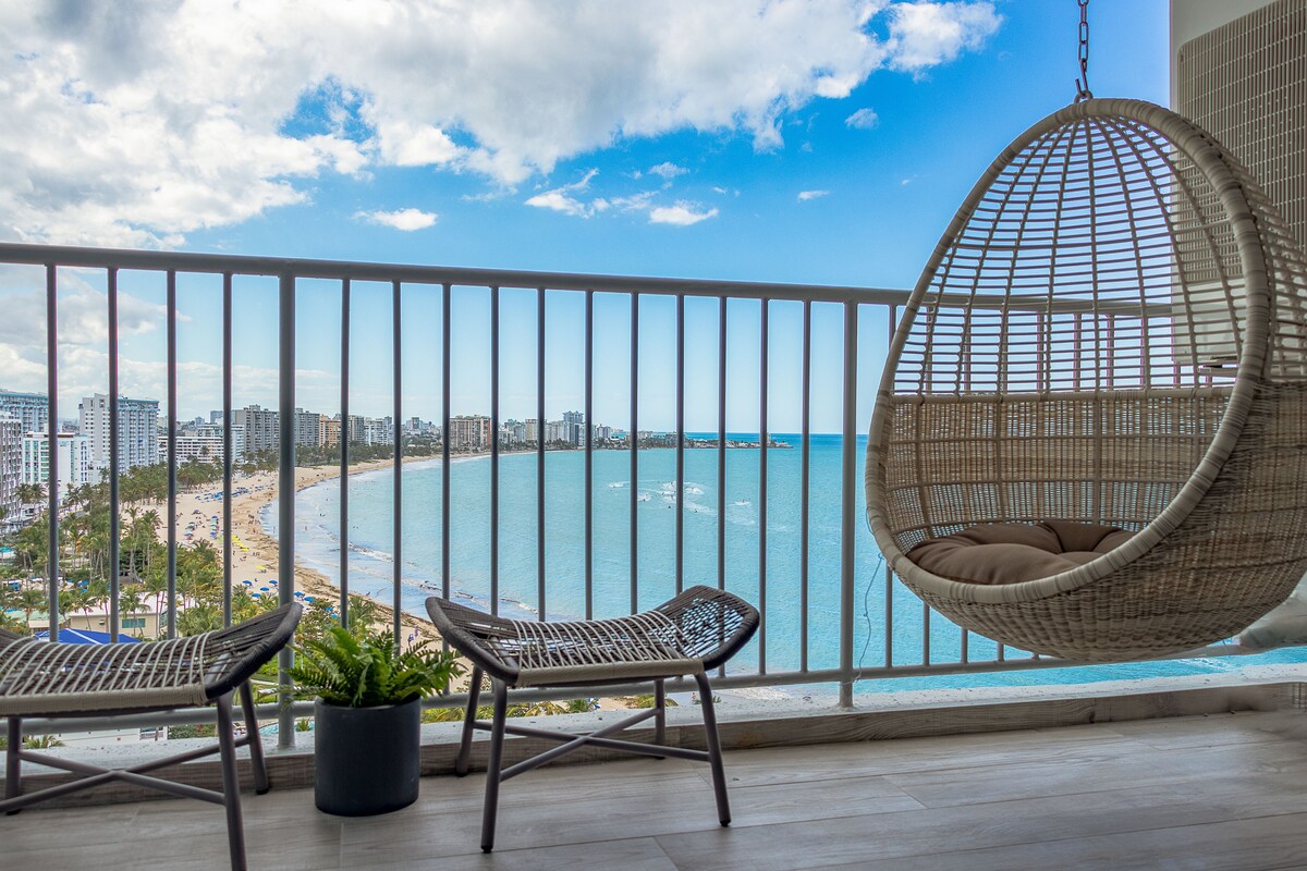 Un balcón con sillas de mimbre, una hamaca colgante y una planta en maceta, con vistas a la playa y al paisaje urbano.