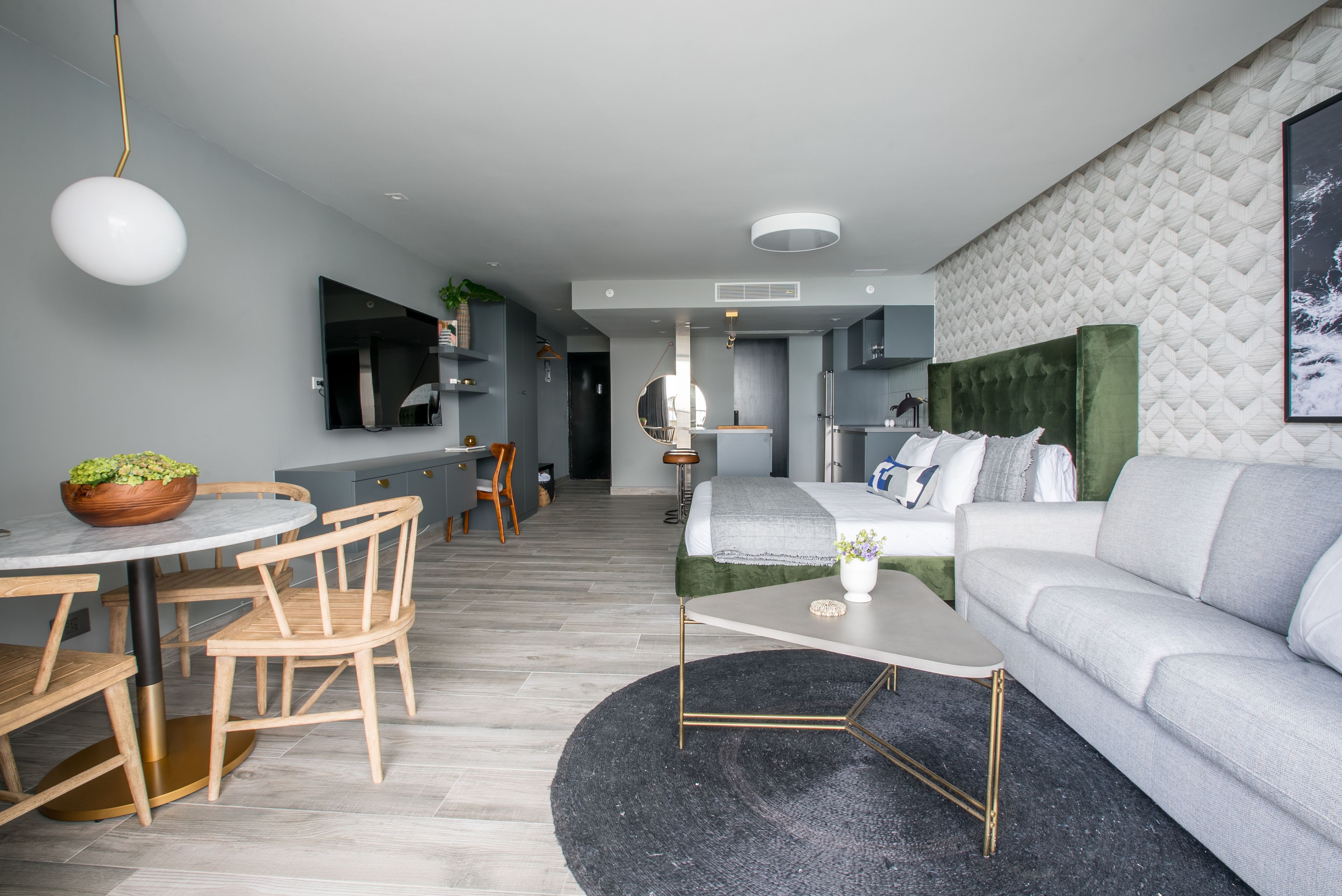 A modern living room with a grey sofa, round coffee table, and wooden dining table with chairs. There is a TV mounted on the wall and a small plant on the table.