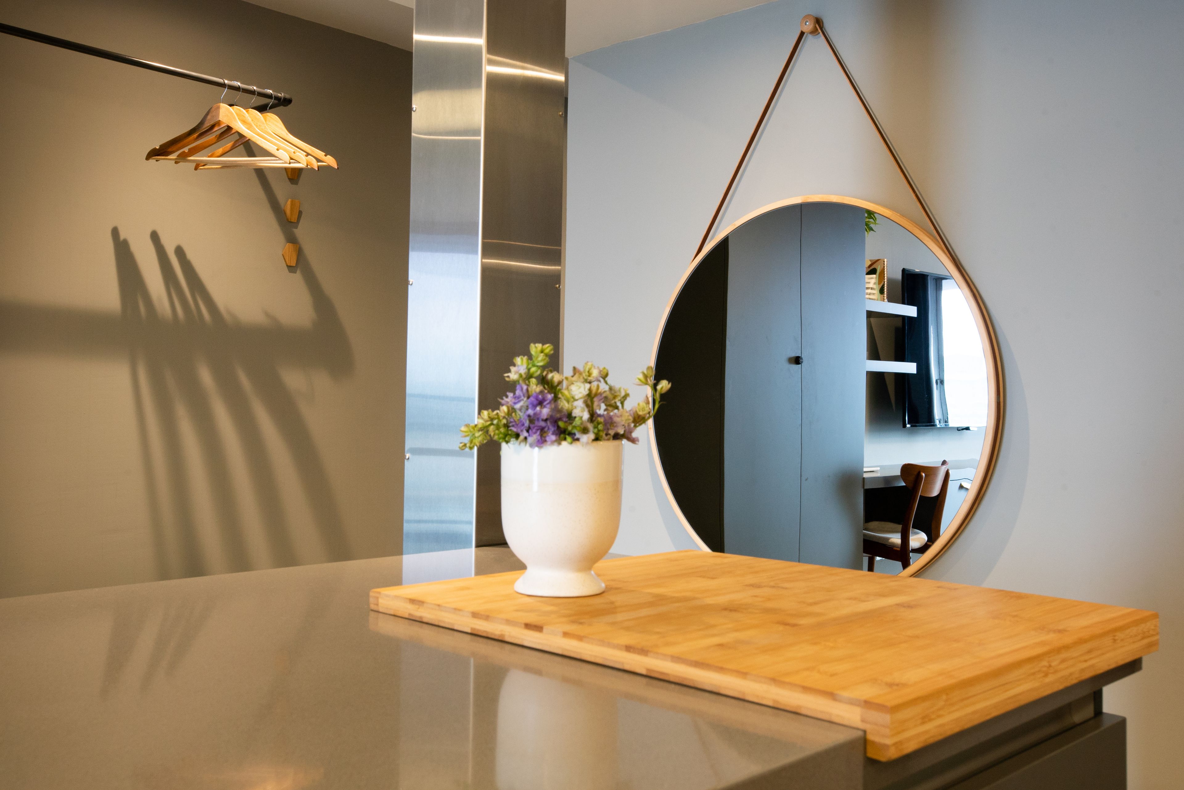 Un mostrador de recepción con superficie de madera, un jarrón blanco con flores, un espejo redondo y un perchero para ropa.