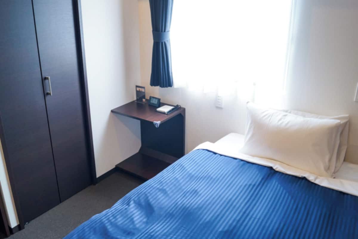 A hotel room with a bed, a nightstand, a desk, a wardrobe, and a window with curtains.