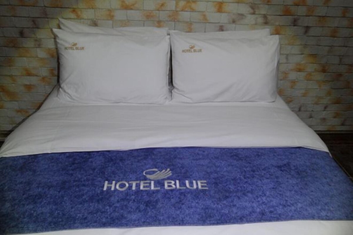 A hotel bed with white linens and pillows, a blue blanket with 'HOTEL BLUE' branding, and a brick-patterned wall behind.