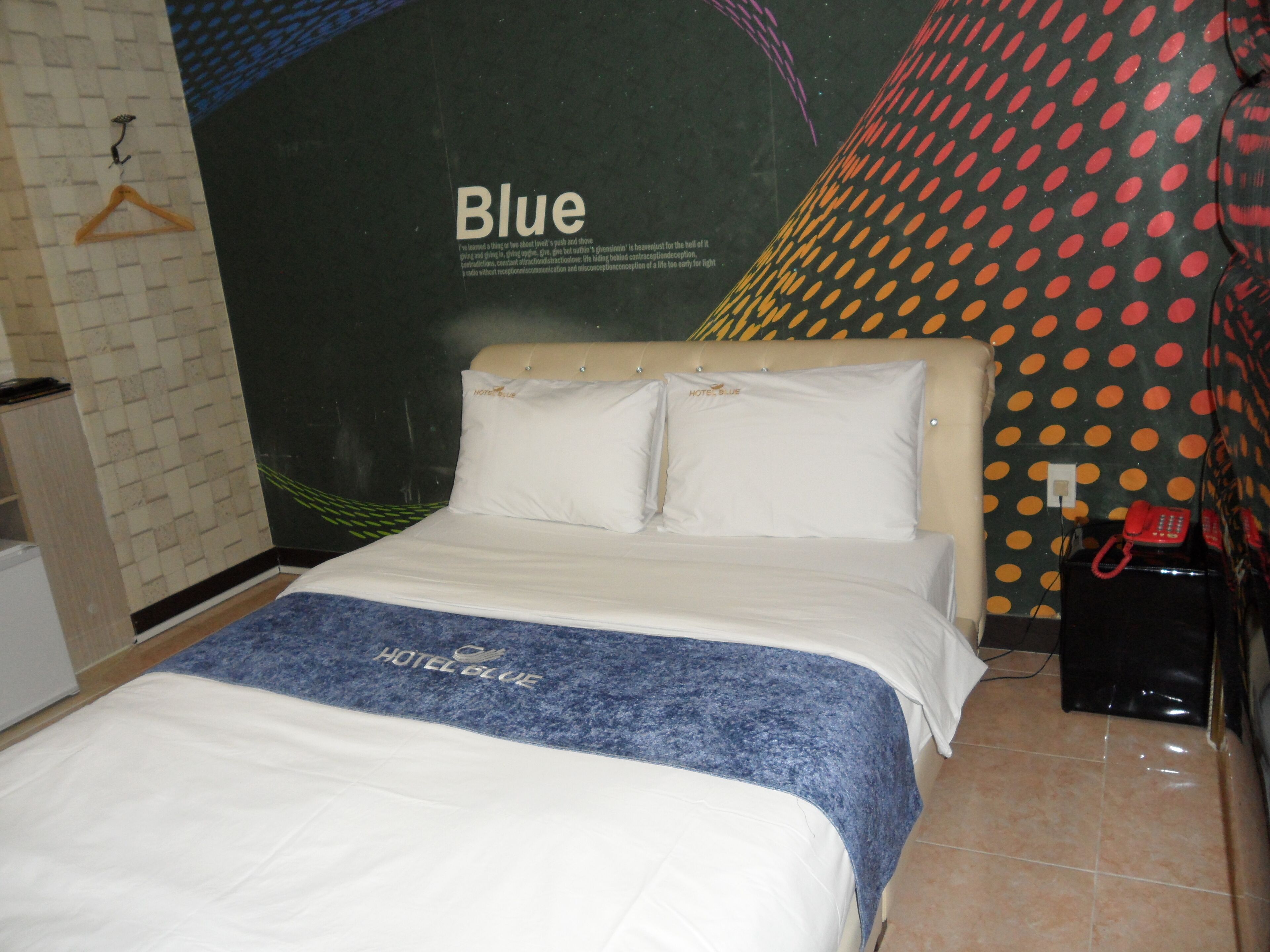 A hotel room with a bed, a wall with a 'Blue' sign, a telephone, and a red object on the floor.