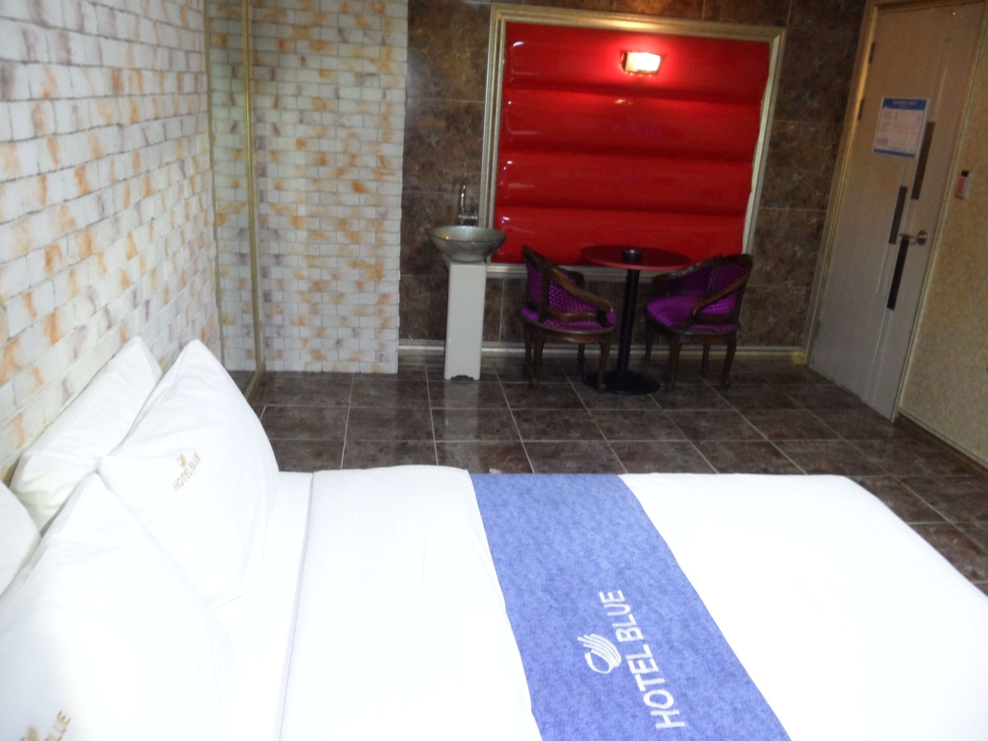 Habitación de hotel con cama, una mesita pequeña con dos sillas, un lavamanos y una puerta roja.