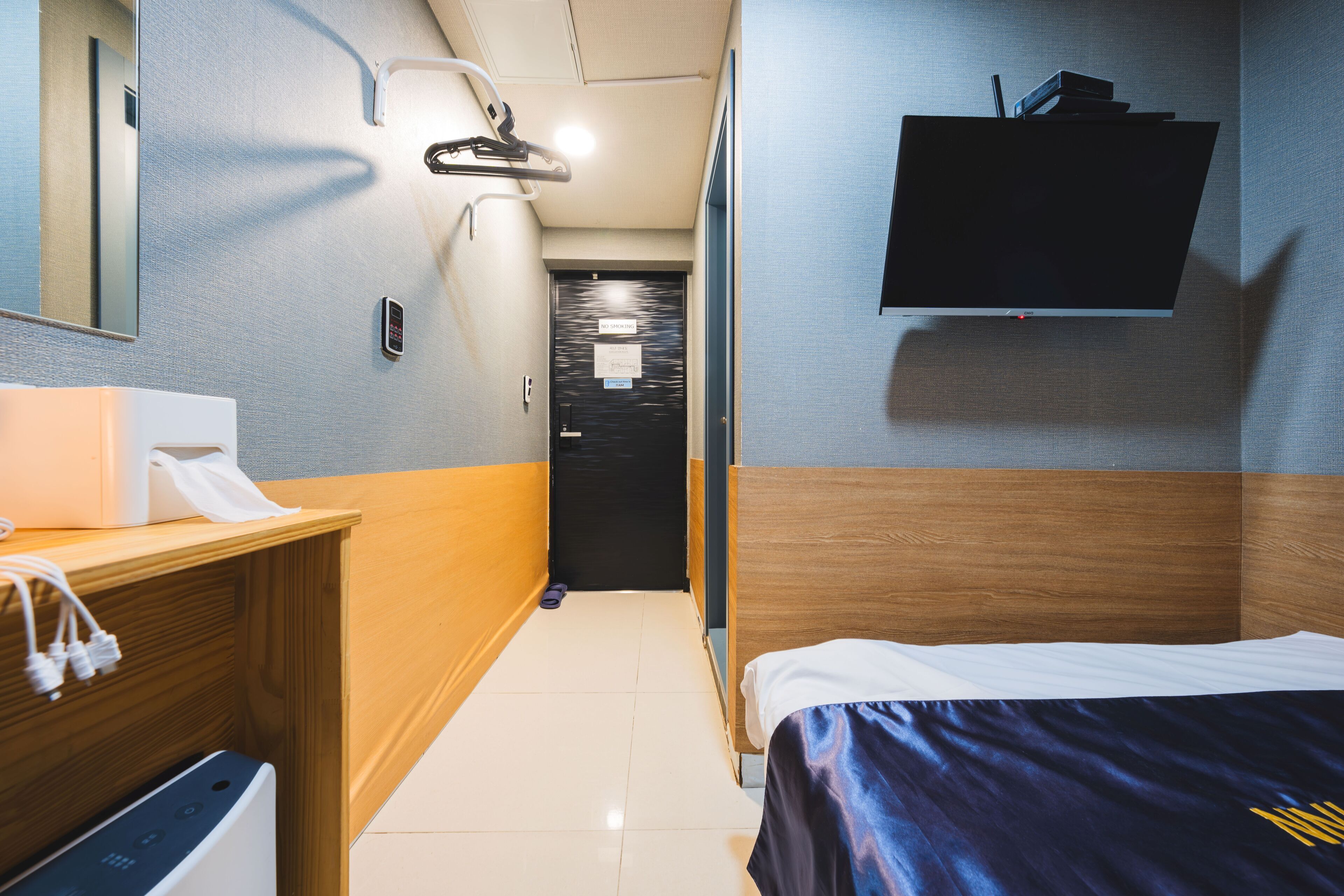 Habitación de hotel con una cama, un aparador de madera, un escritorio con teléfono, un televisor montado en la pared y una puerta que da a un pasillo.