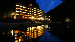 Un hotel de varios pisos con ventanas iluminadas que se reflejan en una masa de agua tranquila, rodeado de vegetación y un paisaje montañoso.