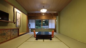 Una habitación de tatami con una mesa baja, cojines naranjas, un televisor en una estantería y vistas a árboles a través de paneles shoji.