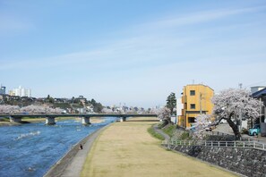 A riverside walkway with a bridge, cherry blossoms, and buildings in the background. - Sararaso (Kanazawa)