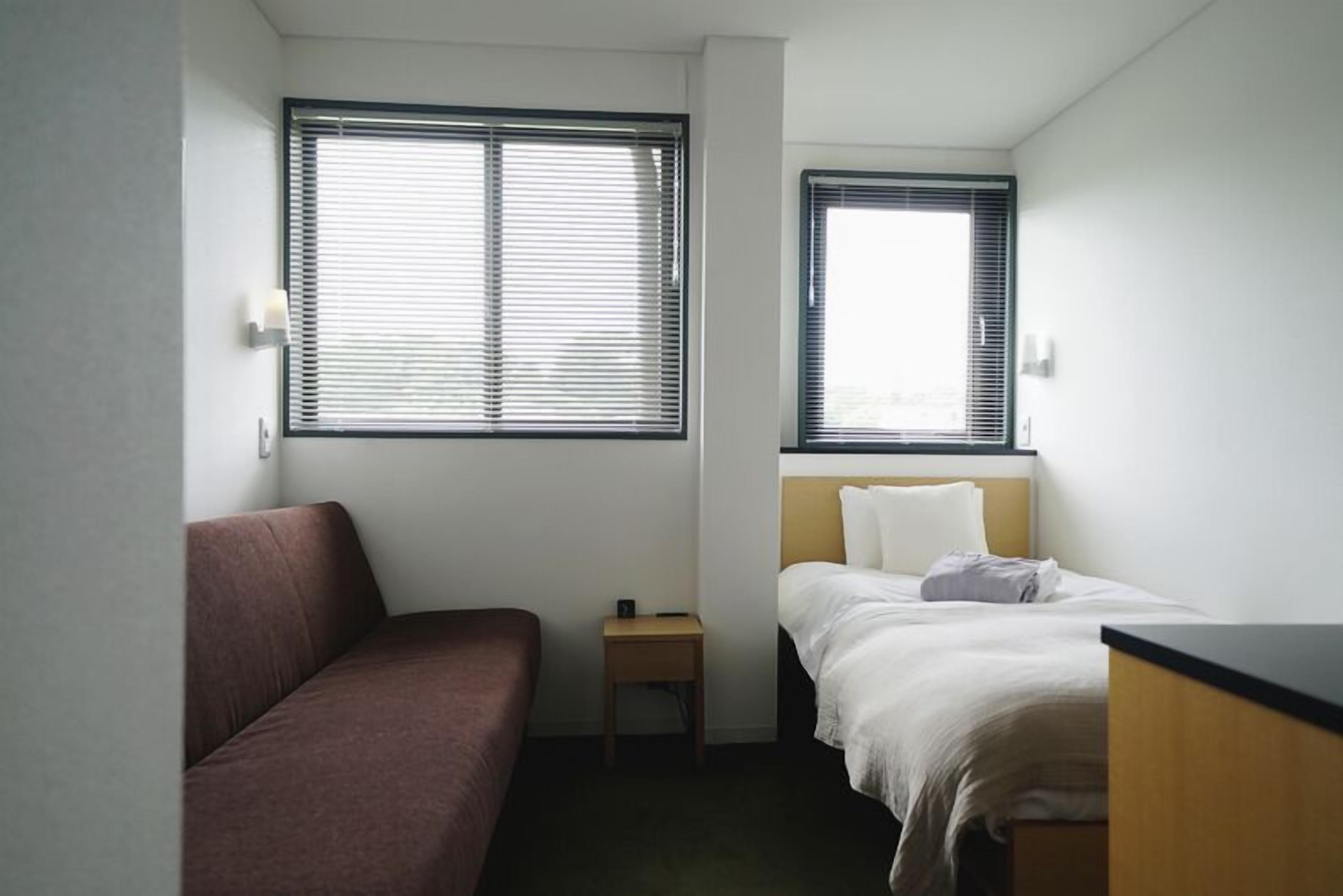 A small, single-room hotel room with a bed, a small bedside table, a couch, and two windows.