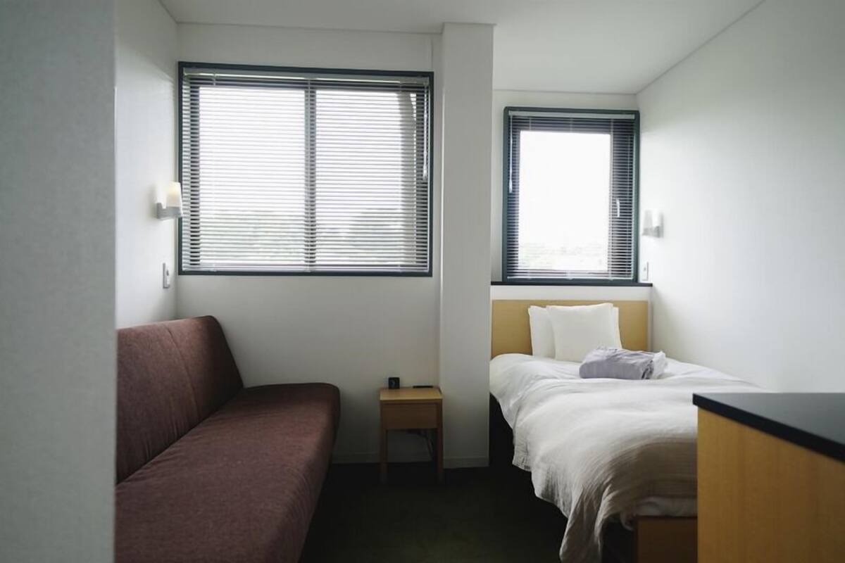 A small, single-room hotel room with a bed, a small bedside table, a couch, and two windows.