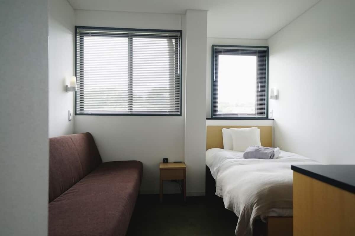 A small, single-room hotel room with a bed, a small bedside table, a couch, and two windows.