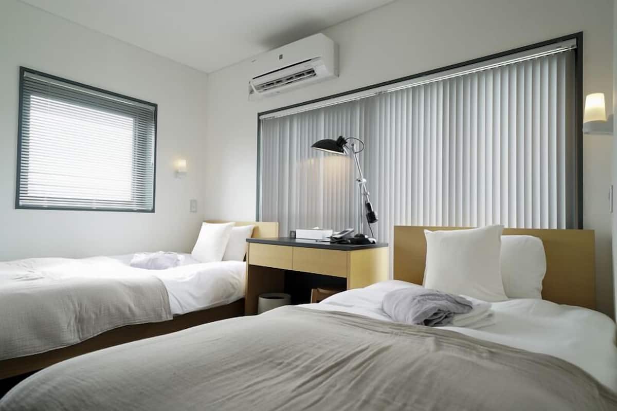 A hotel room with two beds, a desk, a lamp, and an air conditioning unit.
