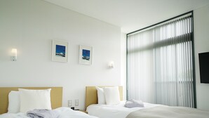 Une chambre d’hôtel avec deux lits, du linge de lit blanc, une grande fenêtre avec des stores et deux tableaux encadrés au mur.