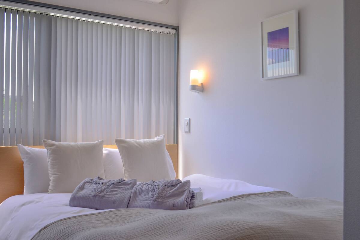 A hotel room with a bed, pillows, a nightstand, a wall-mounted light, and a framed picture.