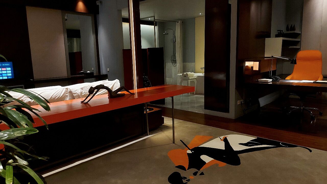 Una habitación de hotel moderna con escritorio, una silla, cama, zona de ducha y un espejo grande.
