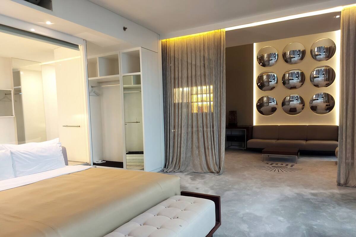 A modern bedroom with a large bed, a walk-in closet, a seating area with a sofa, and a wall adorned with mirrors.