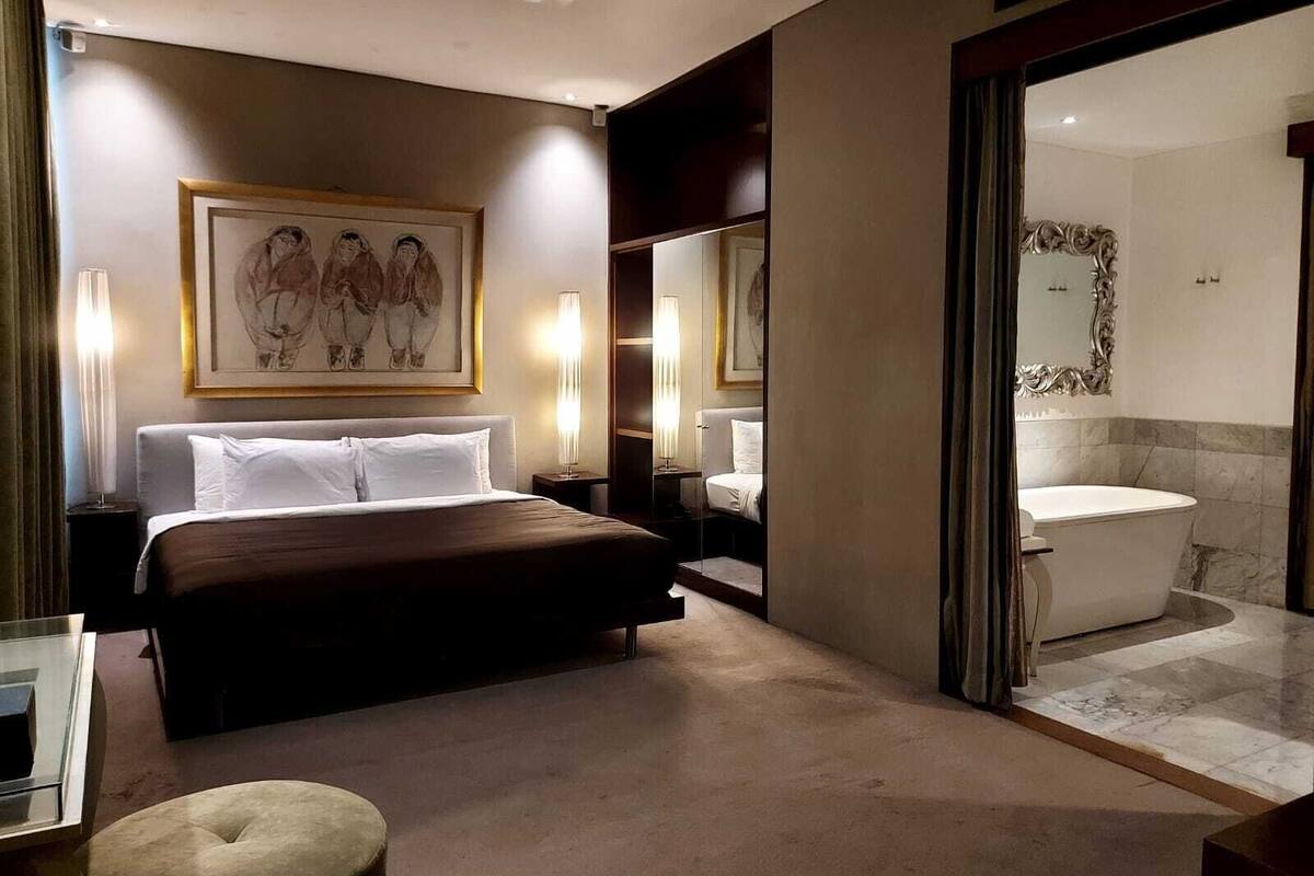 A hotel room with a bed, bedside lamps, a large framed picture, a mirror, and a bathroom with a bathtub.