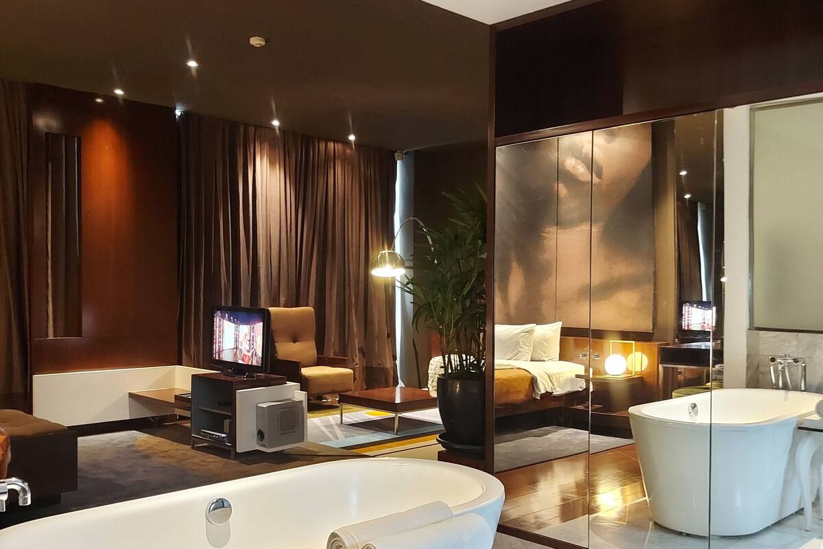 A modern bathroom with a freestanding bathtub, a large mirror, and a seating area with a television.