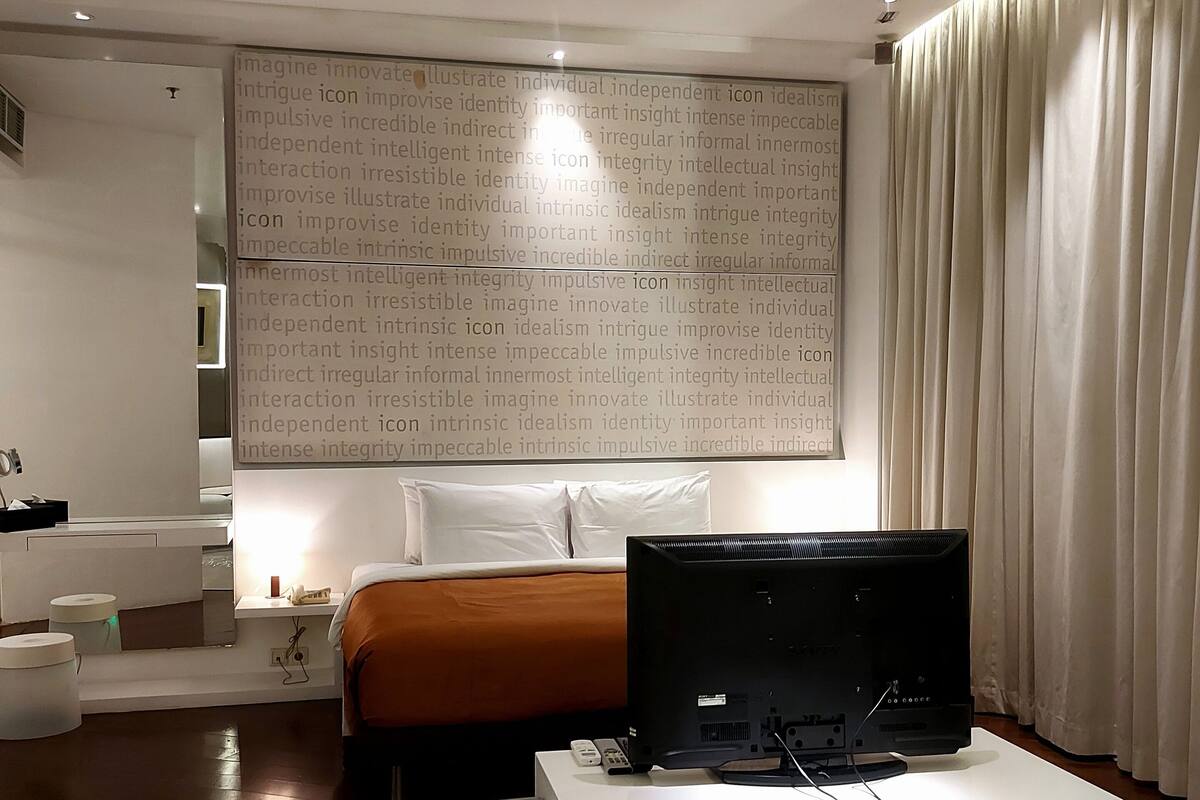 A modern hotel room with a large bed, a flat-screen TV on a white console, a wall with text, and a large window with curtains.
