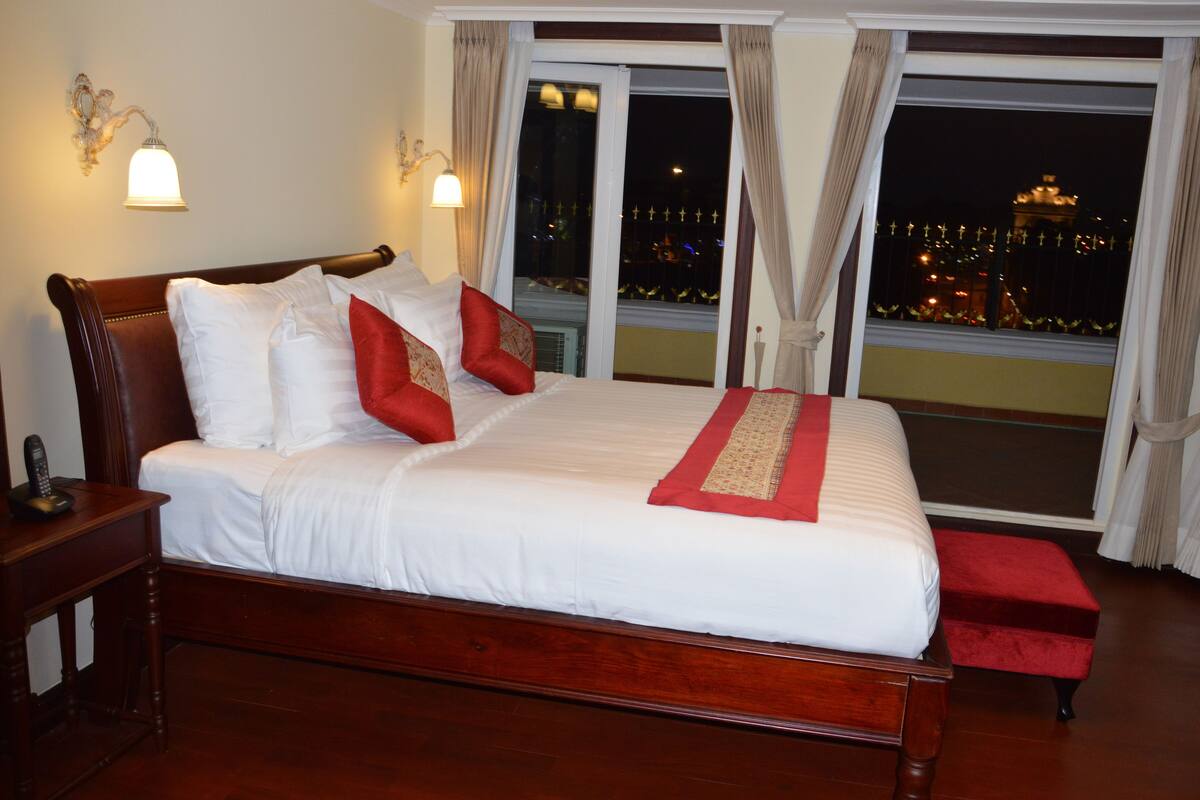 A large bed with white linens and red accents, a wooden nightstand with a telephone, and a view of a cityscape through a large window.